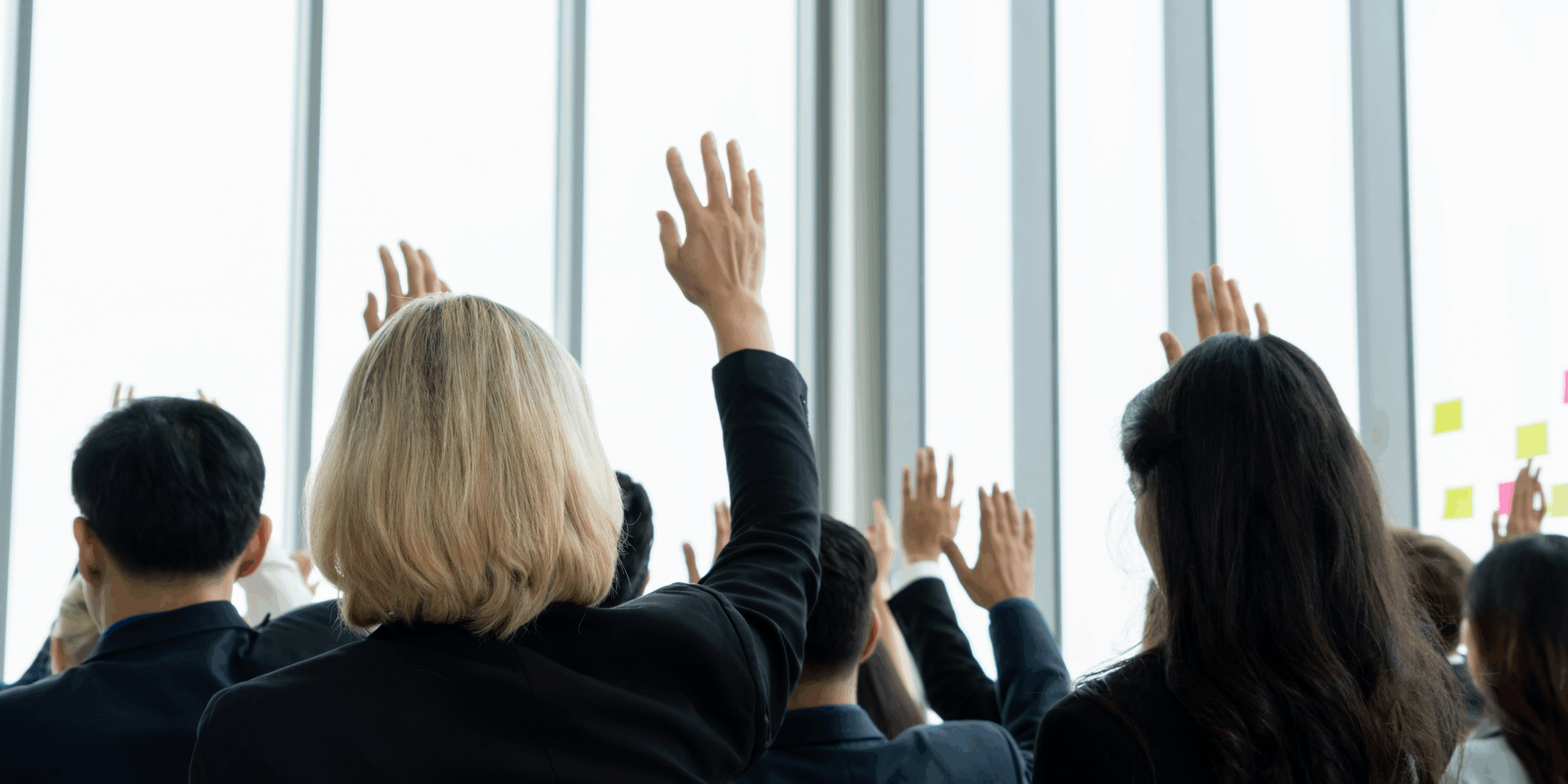 Image depicts a bright, naturally lit room with many professional men and women in suits raising their hands with questions to ask.