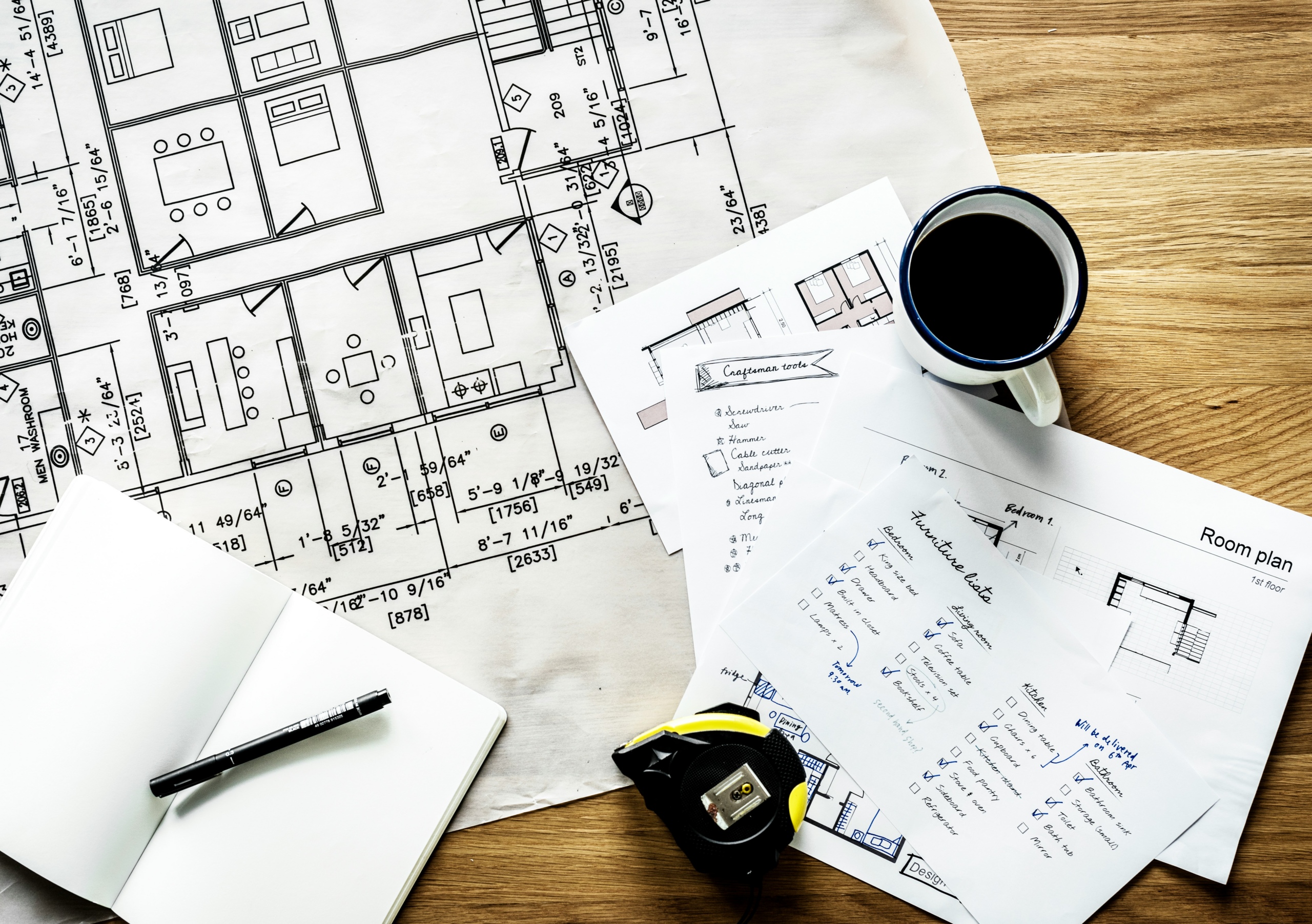 Architectural blueprints, handwritten planning notes, a room layout checklist, a tape measure, and a cup of coffee spread across a wooden work surface — representing the deliberate, step-by-step process of building a custom AI prompt engine designed specifically for your association's workflows and voice.